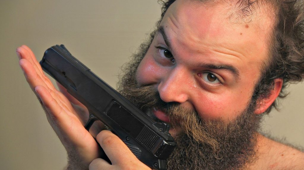 A bearded man mugs for the camera while displaying his pistol | Idiots With Guns: No Backstop Edition [Video] | Featured