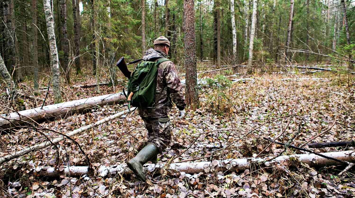 Man the hunter goes through the forest in rubber boots, with a backpack and a gun | Go Bag Guns | Getting Your Firearms In Order When SHTF | Featured